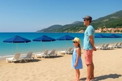 Alleenreizende ouder en dochter samen op een Grieks zandstrand, met turquoise zee, ligbedden en blauwe parasols op de achtergrond.