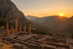 Ruïnes van de tempel van Apollo in Delphi, omgeven door bergen en verlicht door de ondergaande zon.