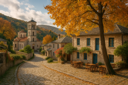 Herfstdorp in Griekenland met kerk, gele bladeren en lege terrasstoelen onder een grote boom in een bergachtig landschap.