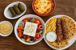 Tafel met traditionele Griekse gerechten waaronder moussaka, souvlaki, dolmades, tzatziki, hummus en Griekse salade op een houten ondergrond.