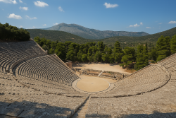 Panoramisch uitzicht op het theater van Epidaurus met stenen zitrijen in halve cirkelvorm, een ronde orchestra en omringende dennenbossen en bergen.
