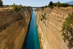 Breedbeeldfoto van het Kanaal van Korinthe met turquoise water tussen hoge, verticale rotswanden en een stalen brug op de achtergrond.