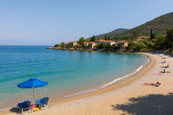 Zand- en kiezelstrand in Agios Nikolaos met blauwe parasols, rustige zee en traditionele huizen tegen een groene heuvel.