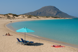 Strand op Elafonisos met licht zand, turquoise zee, ligstoelen en parasol, omgeven door duinen en een groene berg op de achtergrond.