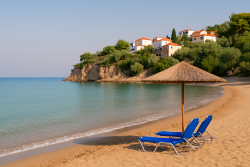 Strand in Ermioni met twee blauwe ligstoelen onder een rieten parasol, kalme zee en witte huizen met rode daken op een groene heuvelrug.