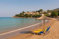 Breed zandstrand in Finikounda met ligstoelen onder een rieten parasol, kalme turquoise zee en witte huizen met rode daken op een beboste helling.