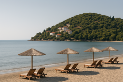 Strand in Gialova met houten ligstoelen onder rieten parasols, kalme zee en groene heuvel met witte huizen op de achtergrond.