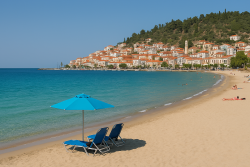 Strand in Gythio met zand, twee ligstoelen onder een blauwe parasol, kalme turquoise zee en pastelkleurige huizen tegen een heuvel.