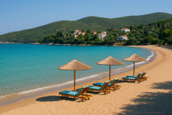 Lang zandstrand in Kalo Nero met ligstoelen en rieten parasols, kalm turquoise water en traditionele huizen op een groene heuvel.