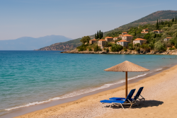 Breed zand- en kiezelstrand in Kardamili met twee blauwe ligstoelen onder een rieten parasol, turquoise zee en stenen huizen op een groene heuvel.
