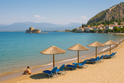 Strand bij Nafplion met kiezels en zand, turquoise zee, uitzicht op het Bourtzi-fort in het water en witte huizen tegen een groene heuvel.