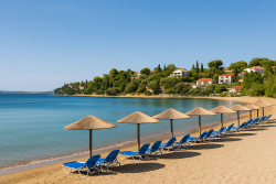 Zandstrand in Porto Heli met rieten parasols, blauwe ligstoelen, turquoise water en villa’s op een groene heuvel aan de overzijde van de baai.