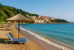 Strand in Pylos met ligstoelen onder rieten parasols, kalm blauw water en witgekalkte huizen tegen een groene heuvelrug.