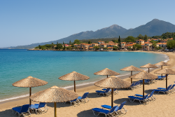 Zandstrand in Stoupa met rijen rieten parasols en blauwe ligstoelen aan een turquoise baai, met huizen en bergen op de achtergrond.
