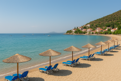 Zandstrand in Tolo met rieten parasols en ligstoelen, kalme turquoise zee en groene eilandjes aan de horizon.