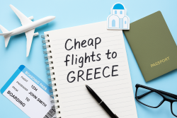 Notitieboek met de tekst “Cheap flights to Greece” naast een modelvliegtuig, paspoort, bril en boardingpass op een lichtblauwe ondergrond.